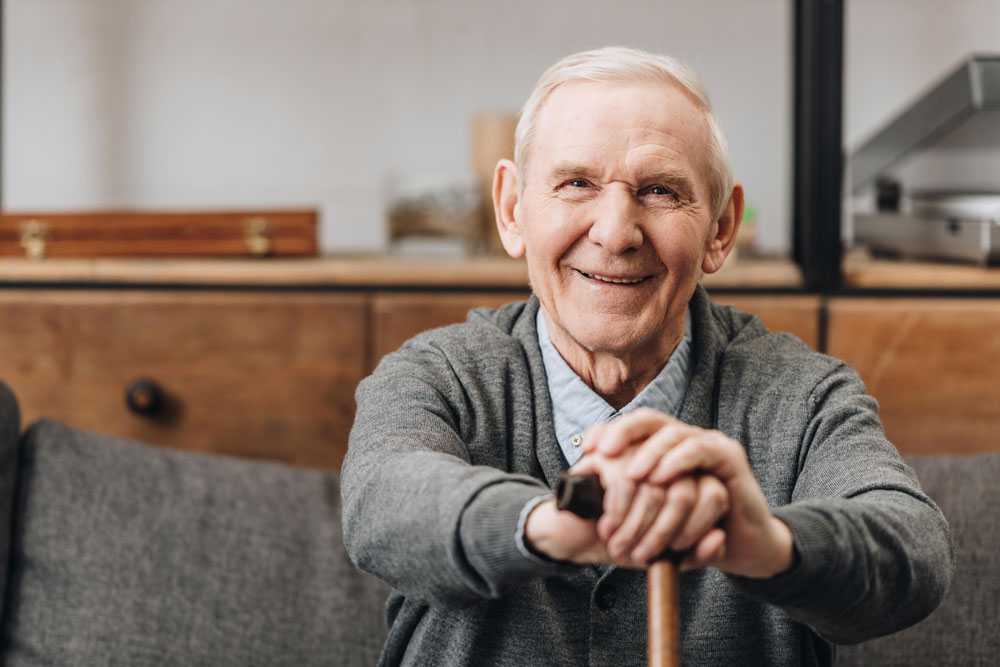 Cheerful old man suffering from loss of balance sitting on sofa smiling and holding walking cane