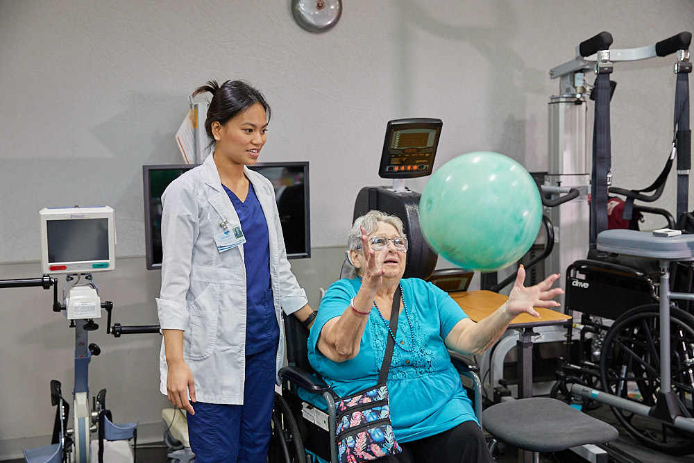 Elderly woman playing with ball sitting on a wheelchair while nurse is standing by her side as part of physical therapy after stroke
