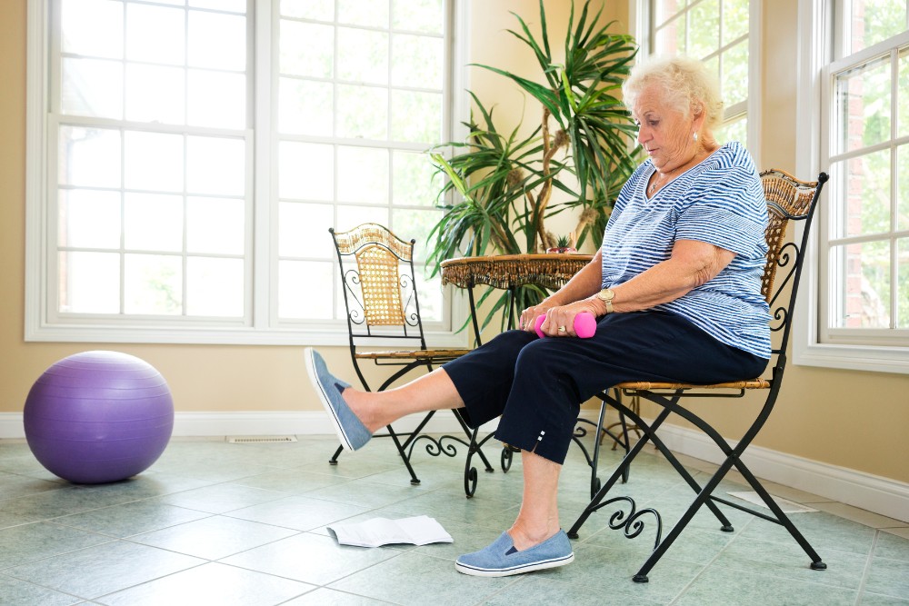 Senior woman exercising while tired, sitting on a chair lifting a small dumbbell and stretching her leg, promoting active aging and fitness.